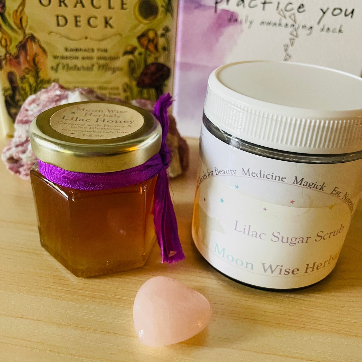 Jar of honey, jar of lilac sugar scrub, and crystal on a wooden surface with 'Green Witch's Oracle Deck' in the background.