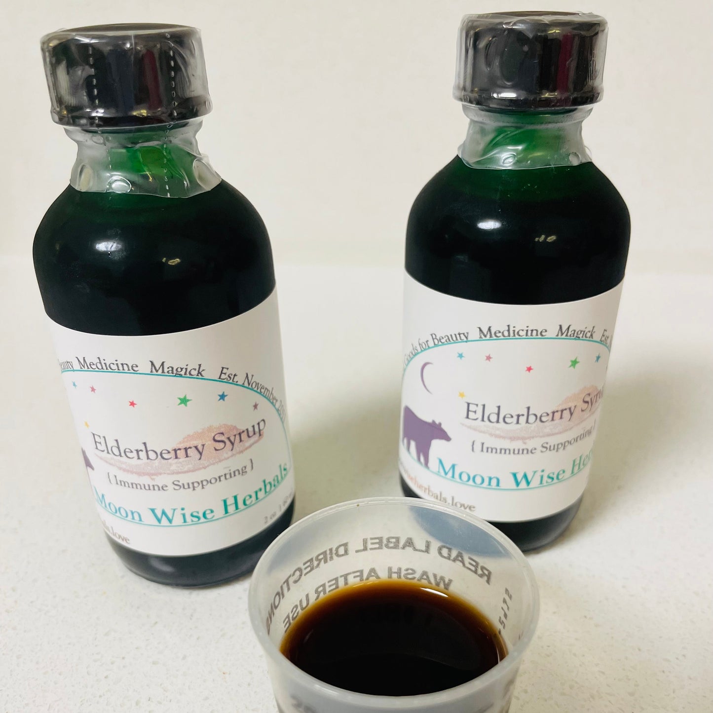 Two bottles of Elderberry Syrup with a small cup filled with the syrup on a white surface.